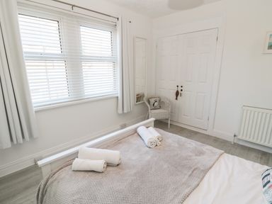 A bedroom with a bed covered with a gray blanket and rolled white towels near a window with blinds and curtains a white chair with a cushion near a white closet door and a wall mirror at 4 Percy Street in Amble