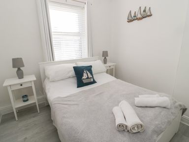 A bedroom with a bed covered by a gray blanket and two rolled towels on top two bedside tables with gray lamps and a window with white blinds and curtains at 4 Percy Street in Amble