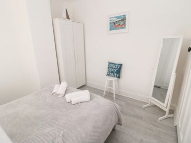 A bedroom with a bed covered in a gray blanket and white towels on it a white wardrobe a white stool with a cushion and a standing mirror at 4 Percy Street in Amble