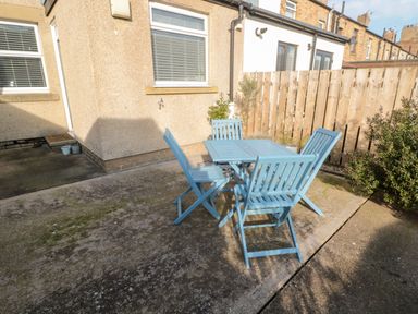 An outdoor patio area with a blue table and four blue chairs beside a beige house and wooden fence at 4 Percy Street in Amble