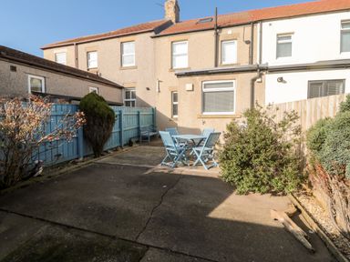 A backyard with a blue wooden table and chairs set next to a beige house with blue fencing and bushes at 4 Percy Street in Amble