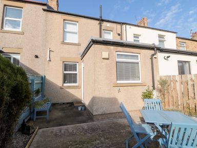 An outdoor patio area with blue wooden chairs and a table next to a beige house with two windows and a blue bench near a blue wooden fence at 4 Percy Street in Amble