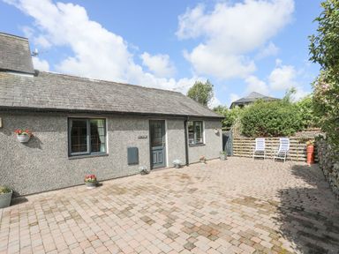 A courtyard with a paved ground and a gray building with two windows and a door with flower pots and two chairs at Cae Ffynnon Star near Gaerwen