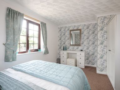 A bedroom with a bed blue bedding window with curtains vanity table mirror and patterned wallpaper at Cae Ffynnon Star near Gaerwen