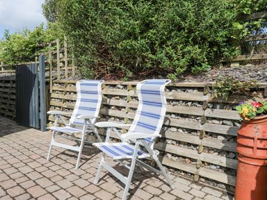 Two striped outdoor chairs on a paved patio beside a stone and wood retaining wall with plants at Cae Ffynnon Star near Gaerwen