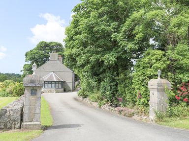 A driveway entrance with stone pillars and lanterns leading to a house surrounded by trees and greenery at Cae Ffynnon in Star near Gaerwen
