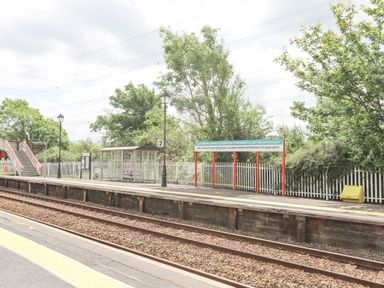 A railway platform with a sign displaying a long place name a glass shelter trees and a pedestrian bridge at Cae Ffynnon Star near Gaerwen
