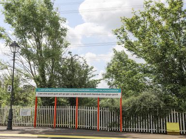 A railway platform with a sign showing a long place name and green trees behind a white fence at Cae Ffynnon in Star near Gaerwen