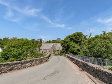 A road crossing a stone bridge with trees on both sides and a stone building in the background at Ty Eiddew Ivy House in Llanystumdwy near Criccieth
