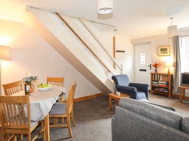 A living room with a dining table and chairs under stairs a blue armchair a gray sofa bookshelf and a door at Ty Eiddew Ivy House in Llanystumdwy near Criccieth