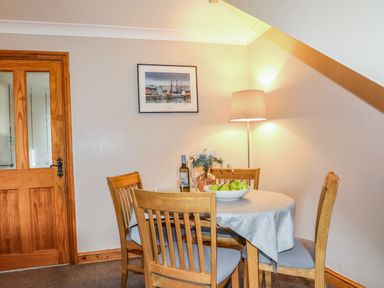 A dining area with a round table covered by a tablecloth surrounded by four wooden chairs a bowl of green apples a bottle of wine and a floor lamp on the side at Ty Eiddew in Llanystumdwy near Criccieth