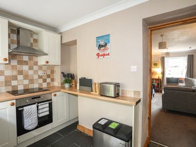 A kitchen with a stove, oven, toaster, utensils, and bins next to an open doorway leading to a living room at Ty Eiddew (Ivy House) Llanystumdwy near Criccieth