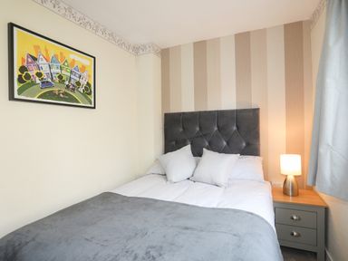 A bed with a gray headboard two pillows a bedside table with a lamp and a colorful framed picture on the wall at Ty Eiddew Ivy House in Llanystumdwy near Criccieth