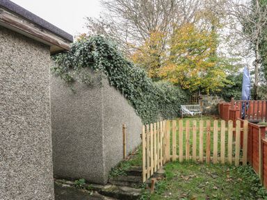 A small garden area with a wooden gate, steps, and a bench surrounded by trees and fencing at Ty Eiddew Ivy House in Llanystumdwy near Criccieth