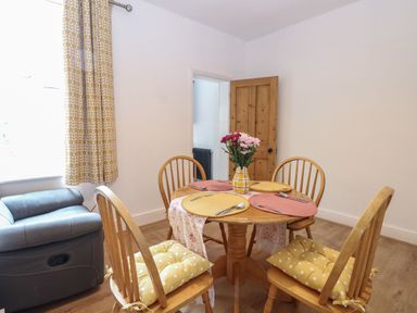 A dining room with a round wooden table set with placemats and utensils four wooden chairs with yellow cushions a vase with flowers a gray armchair near a window with patterned yellow curtains and a wooden door open at Sole Bay Cottage in Lowestoft