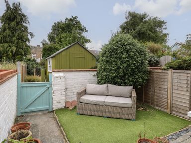 A small garden area with a gray cushioned wicker sofa on artificial grass next to a green shed with a blue gate and white brick and wooden fences