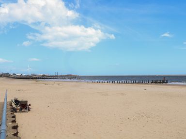 A sandy beach with two people sitting on chairs near a metal railing by the sea at Sole Bay Cottage in Lowestoft