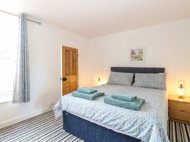 A bedroom with a double bed two folded towels bedside tables lamps a wooden door window with curtains and striped carpet at Sole Bay Cottage in Lowestoft