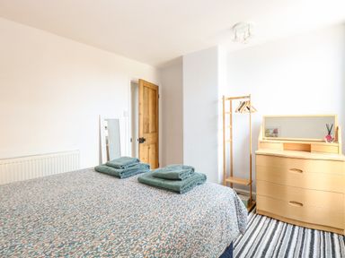 A bedroom with a bed covered in patterned bedding and folded towels on top next to a wooden door a standing mirror a wooden clothes rack with hangers a wooden dresser with a mirror and a striped rug on the floor at Sole Bay Cottage in Lowestoft