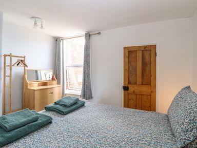A bedroom with a patterned bedspread two folded towels on the bed a wooden door a wooden dresser with mirror a small clothes rack and a window with curtains at Sole Bay Cottage in Lowestoft