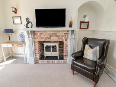 A room with a brick fireplace with a stove inside a black leather armchair with a pillow a small wooden table with a lamp and a blue and white vase a mounted television and decorative items at 9 Station Cottages in Belford