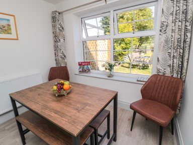A dining area with a wooden table, two brown chairs, a bowl of fruit on the table, a window with patterned curtains, and a view of a garden at 9 Station Cottages in Belford
