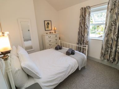 A bedroom with a white metal bed frame a white chest of drawers a mirror and a window with patterned curtains at 9 Station Cottages in Belford