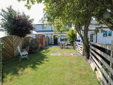 A garden with a white house in the background grass a wooden fence a white metal bench a table with four chairs and trees at 9 Station Cottages in Belford