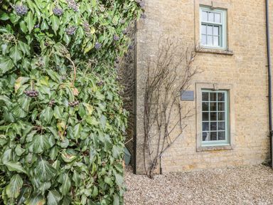 Side of a stone building with two windows and climbing plants next to a leafy shrub at Barton Cottage in Bourton-On-The-Water