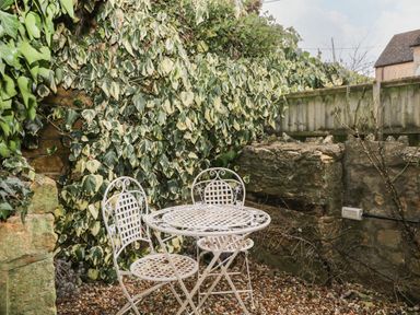 A small outdoor seating area with a metal table and two chairs surrounded by stone walls and dense climbing plants at Barton Cottage in Bourton-On-The-Water