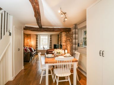 A dining area with a wooden table set for four and white chairs in an open plan living room with exposed wooden beams and stone walls at Barton Cottage in Bourton-On-The-Water