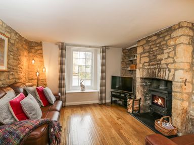 A living room with a brown leather sofa with cushions a window with plaid curtains a television and a stone fireplace with a wood burner at Barton Cottage in Bourton-On-The-Water