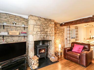 A living room with a stone fireplace wood logs a brown leather armchair with a pink cushion and a television on a stand at Barton Cottage in Bourton-On-The-Water