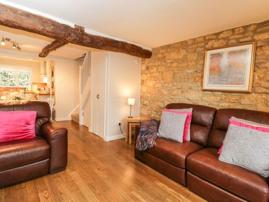A living room with brown leather sofas with pink and gray cushions a stone wall with framed artwork a wooden floor and a lamp on a small table at Barton Cottage in Bourton-On-The-Water
