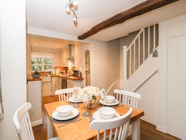 A dining area with a wooden table set for four next to a kitchen with white cabinets and a staircase with white railing at Barton Cottage in Bourton-On-The-Water