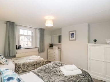 A bedroom with two single beds dolls with blankets a window with curtains a chest of drawers and a wardrobe at Barton Cottage in Bourton-On-The-Water