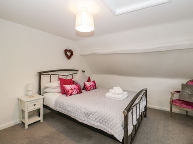 A bedroom with a metal bed frame gray quilt and pink pillows a bedside table with a lamp and an armchair with a cushion at Barton Cottage in Bourton-On-The-Water