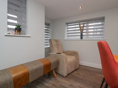 A sitting area with a beige armchair, a bench with brown and gray upholstery, a red chair, and windows with striped blinds at Crag View Cottage in Stainton with Adgarley near Dalton-In-Furness