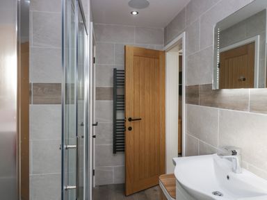 A bathroom with a wooden door a glass shower door a white sink a mirror and tiled walls at Crag View Cottage in Stainton with Adgarley near Dalton-In-Furness