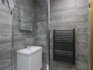 A bathroom with gray tiled walls a white sink with cabinet a mirror and a black towel radiator at Crag View Cottage in Stainton with Adgarley near Dalton-In-Furness