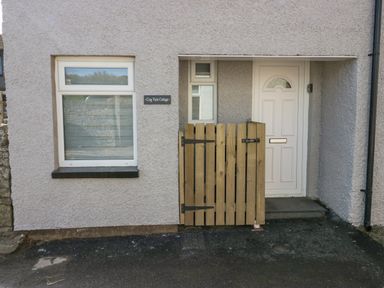 A white front door with a small wooden gate and a window on a textured exterior wall at Crag View Cottage in Stainton with Adgarley near Dalton-In-Furness