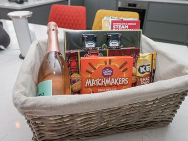 A wicker basket with a bottle of sparkling wine biscuits snacks and jam jars on a kitchen counter at Crag View Cottage in Stainton with Adgarley near Dalton-In-Furness