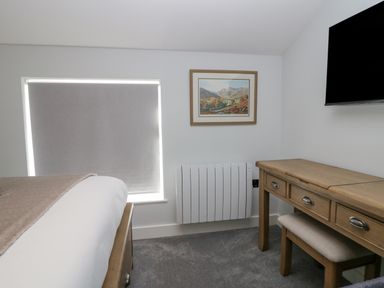 A bedroom with a bed a window with a blind a wall heater a wooden desk with a stool a framed picture and a mounted television at Crag View Cottage in Stainton with Adgarley near Dalton-In-Furness