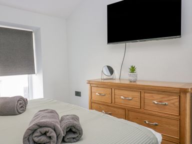 A bedroom with a wooden dresser a mounted TV a small round mirror and a plant on the dresser with rolled towels on the bed at Crag View Cottage in Stainton with Adgarley near Dalton-In-Furness