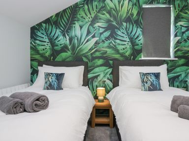 A bedroom with two single beds with white bedding and rolled towels on each bed a small wooden nightstand with a lamp between the beds and tropical leaf wallpaper on the wall at Crag View Cottage in Stainton with Adgarley near Dalton-In-Furness