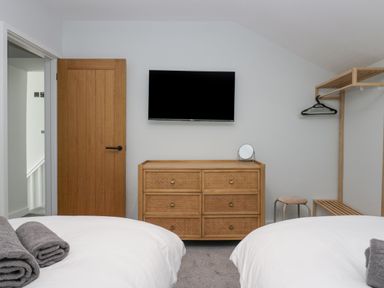 A bedroom with two beds towels on beds a wooden door wooden dresser with a mirror wall mounted tv and wooden clothes rack at Crag View Cottage in Stainton with Adgarley near Dalton-In-Furness