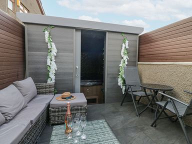 An outdoor seating area with a grey sofa set, a table with a bottle and glasses, and a small shed decorated with white flowers at Crag View Cottage in Stainton with Adgarley near Dalton-In-Furness