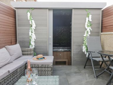 An outdoor seating area with a wicker sofa and ottoman with cushions a glass table with a bottle and glasses a chair and a shed with sliding door decorated with white flowers at Crag View Cottage in Stainton with Adgarley near Dalton-In-Furness
