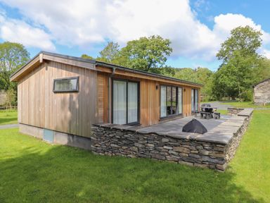 A wooden house with large windows and stone wall patio in a grassy area at Corning Mill View in Haverthwaite
