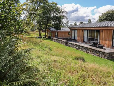 A wooden house with stone terrace and outdoor seating surrounded by grass and trees at Corning Mill View in Haverthwaite